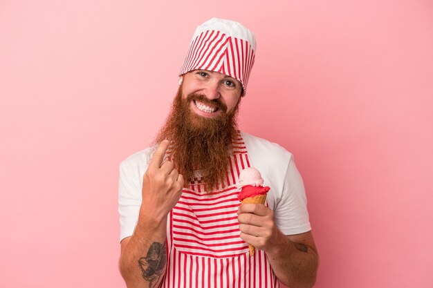 Joven pelirrojo caucásico con barba larga sosteniendo un helado aislado sobre fondo rosa apuntando con el dedo como si invitara a acercarse.