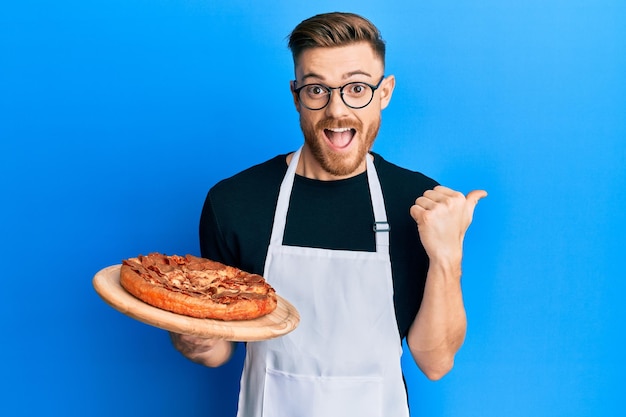 Joven pelirrojo con delantal de camarero sosteniendo pizza apuntando con el pulgar hacia el lado sonriendo feliz con la boca abierta