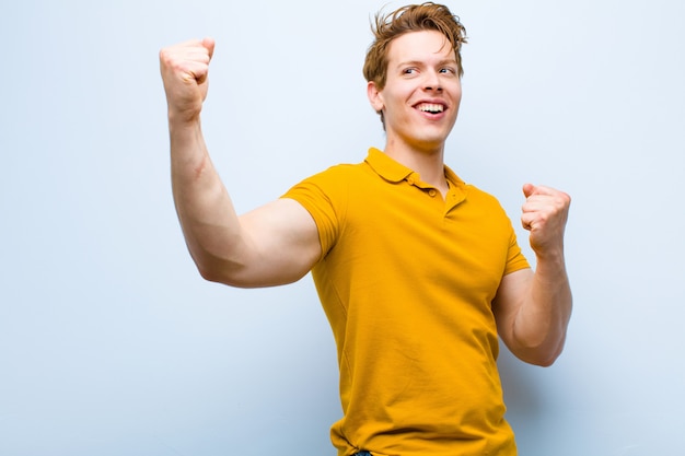 Joven pelirrojo gritando triunfante, luciendo emocionado, feliz y sorprendido ganador, celebrando contra la pared azul