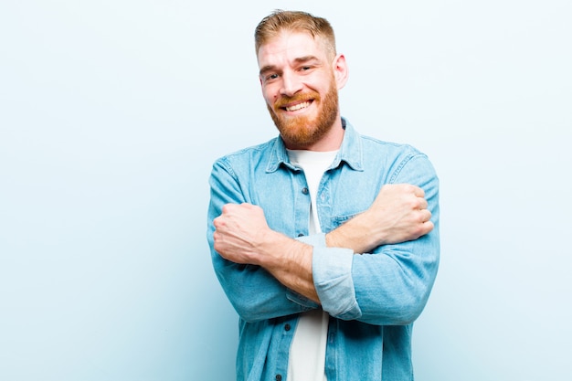 Joven pelirrojo sonriendo alegremente y celebrando, con los puños cerrados y los brazos cruzados, sintiéndose feliz y positivo contra la suave pared azul