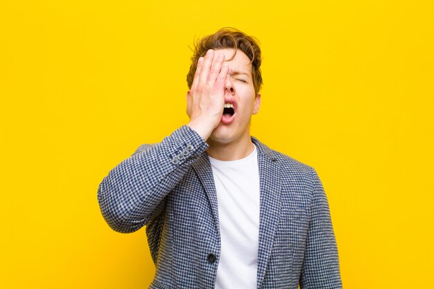 Joven pelirrojo con sueño, aburrido y bostezo, con dolor de cabeza y una mano cubriendo la mitad de la cara