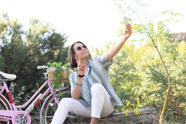 Joven de pelo largo tomándose una selfie con su bicicleta moderna retro rosa en la parte de atrás mientras está en el parque