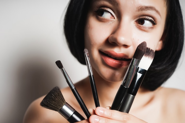 Una joven de pelo negro sosteniendo pinceles de maquillaje, mirando a la izquierda con interés, sonriendo. Aislado sobre fondo blanco