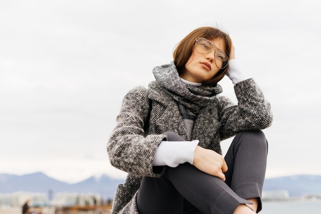 Una joven de pelo oscuro con un abrigo gris y gafas está sentada al aire libre, pensando en algo