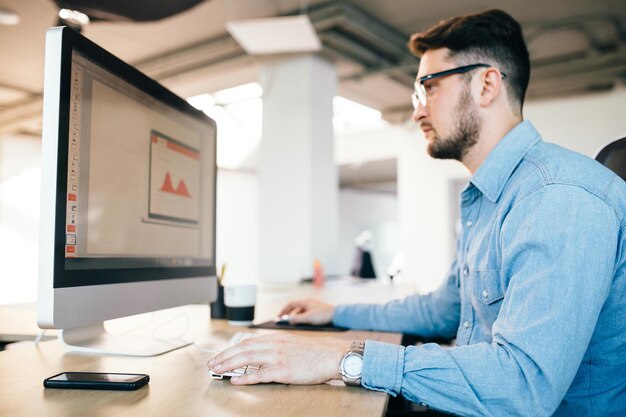 Joven de pelo oscuro en glasess y una camisa azul está trabajando con una computadora en su escritorio,