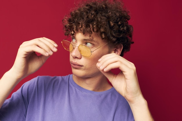 Joven de pelo rizado con gafas amarillas, camiseta morada, emociones, fondo rojo.