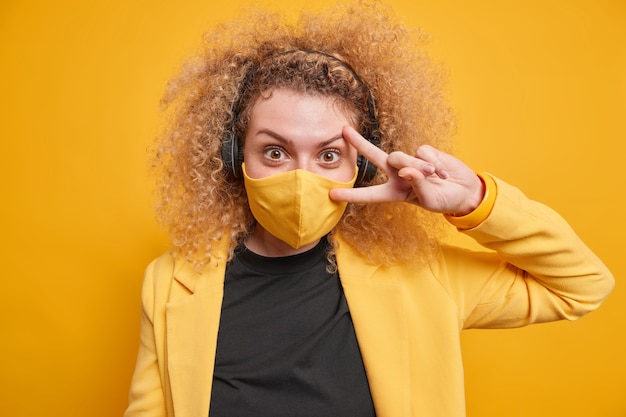 Una joven de pelo rizado positivo hace un gesto de paz sobre el ojo y usa una máscara protectora mientras visita lugares públicos vestida con ropa formal elegante y escucha música a través de auriculares. Vida durante la cuarentena