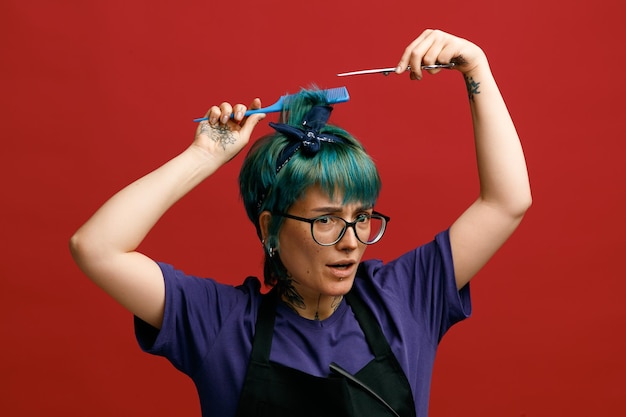 Una joven peluquera frunciendo el ceño con anteojos uniformes y una banda para la cabeza mirando a un lado peinándose el cabello sosteniendo unas tijeras sobre la cabeza haciéndose un corte de pelo con una pinza para el cabello en su delantal