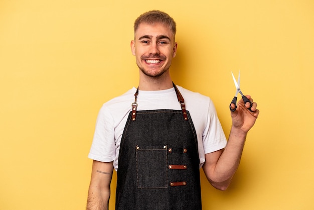 Joven peluquero caucásico sosteniendo tijeras aisladas de fondo amarillo feliz, sonriente y alegre.