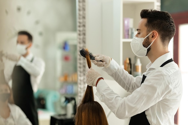 Joven peluquero cepillando el cabello del cliente mientras usa una máscara protectora en un salón