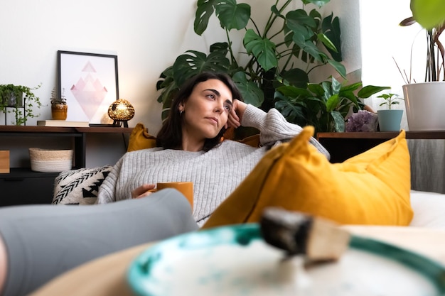 Una joven pensativa en casa tomando té sentada en un sofá mirando por la ventana. Concepto de estilo de vida.