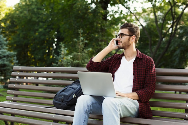 Joven pensativo hablando por móvil, trabajando en una laptop, sentado en el parque. Concepto de tecnología, comunicación, educación y trabajo remoto, espacio de copia