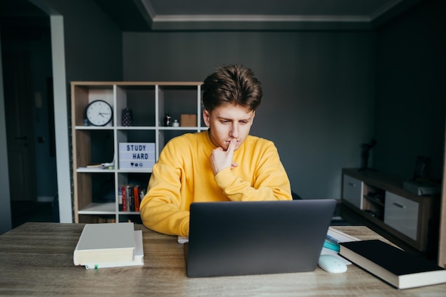 Un joven pensativo con una sudadera amarilla se sienta en casa en una mesa en la habitación y usa una computadora portátil mirando pensativamente la pantalla Estudiante estudiando en casa usando una computadora portátil Cuarentena