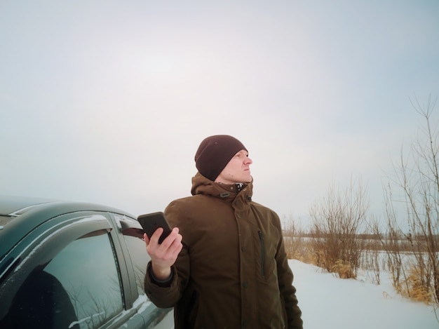 Foto un joven se perdió en un automóvil en invierno y está tratando de alcanzar la red móvil después de salir del automóvil. concepto de viaje por carretera de invierno, búsqueda de carreteras, navegación.