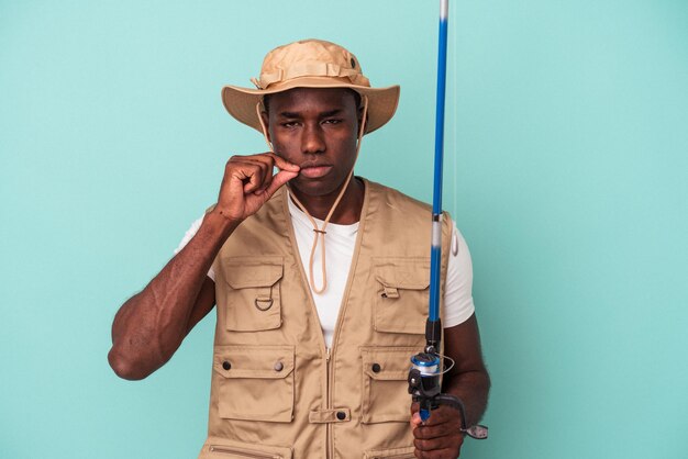 Joven pescador afroamericano sosteniendo una caña aislada de fondo azul con los dedos en los labios guardando un secreto.