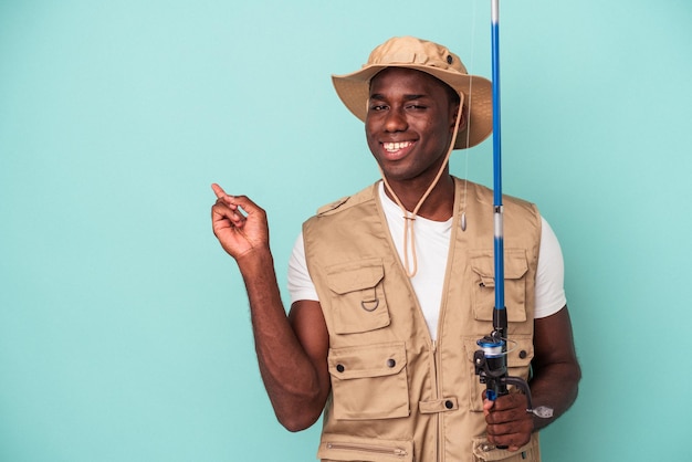 Joven pescador afroamericano sosteniendo una caña aislada de fondo azul sonriendo y señalando a un lado, mostrando algo en el espacio en blanco.