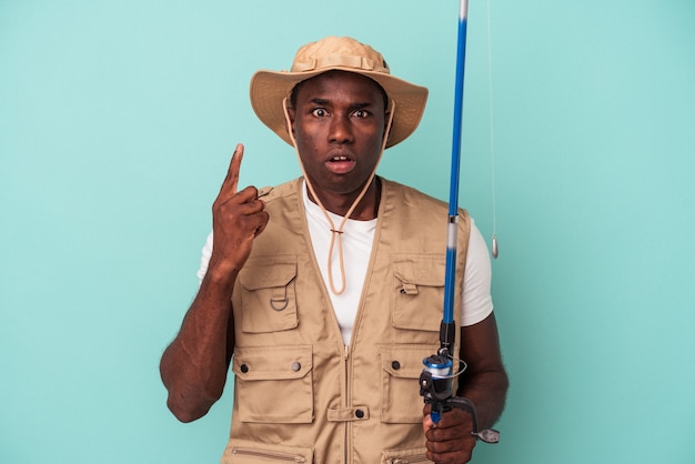 Joven pescador afroamericano sosteniendo la caña aislada sobre fondo azul con una gran idea, concepto de creatividad.