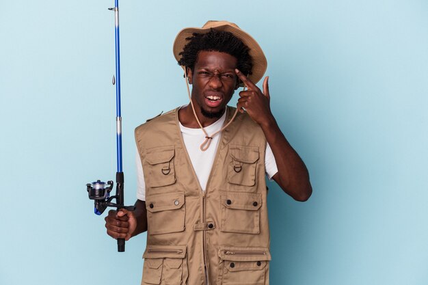 Joven pescador afroamericano sosteniendo una caña aislada sobre fondo azul mostrando un gesto de decepción con el dedo índice.