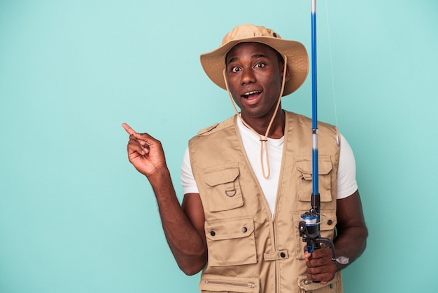 Joven pescador afroamericano sosteniendo la varilla aislado sobre fondo azul apuntando hacia el lado