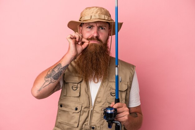 Joven pescador de jengibre caucásico con barba larga sosteniendo una caña aislada sobre fondo rosa con los dedos en los labios guardando un secreto.