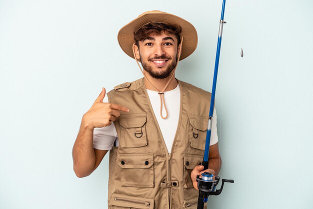 Joven pescador de raza mixta sosteniendo una caña aislada sobre fondo azul persona apuntando con la mano a un espacio de copia de camisa, orgulloso y seguro