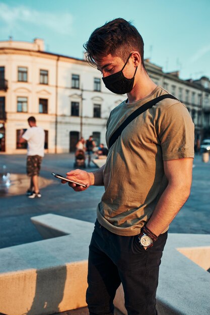 Foto un joven de pie en el centro de la ciudad por la noche usando un teléfono inteligente con la máscara facial