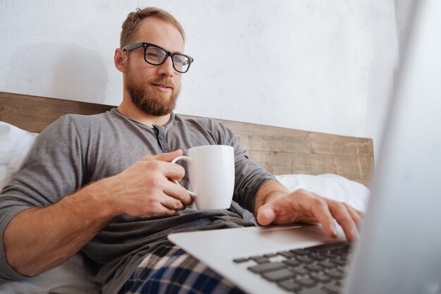 Joven en pijama disfrutando del proceso de trabajo mientras sostiene una taza de café y mira en la pantalla de una computadora portátil en la cama
