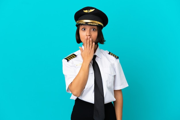 Joven piloto de avión sobre fondo azul aislado cubriendo la boca con la mano