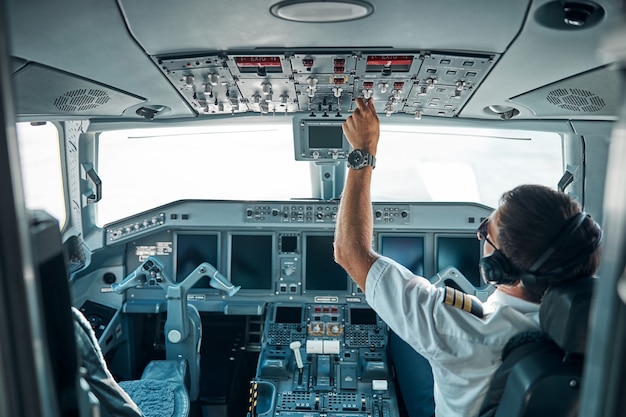 Joven piloto en uniforme y dispositivos está sentado al control en la cabina y usa conmutadores para volar un avión