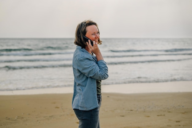 Una joven en la playa cerca del océano en primavera al atardecer hablando por teléfono con una sonrisa