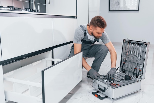 Foto joven plomero profesional con uniforme gris usando equipo en la cocina y preparándose para el trabajo