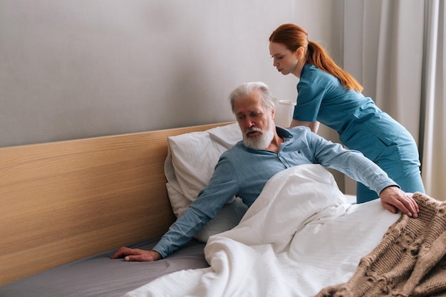 Una joven y positiva cuidadora doméstica que ayuda a un paciente varón adulto mayor a acostarse en la cama en el dormitorio del hogar de ancianos