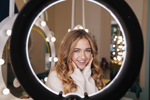 joven positiva linda chica rubia alegre en el interior de un salón de belleza mirando a través de la lámpara de luz de anillo.