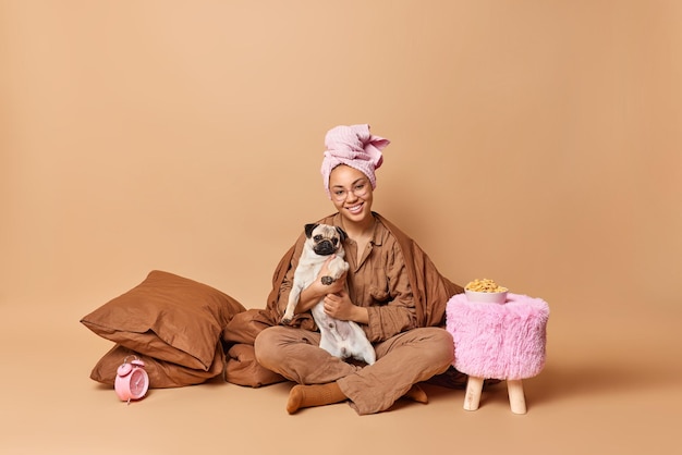 Una joven positiva se sienta con las piernas cruzadas con su mascota favorita, usa pijama y una toalla envuelta en la cabeza, posa bajo una manta, se despierta de buen humor aislada sobre el fondo marrón del estudio. Es hora de dormir.