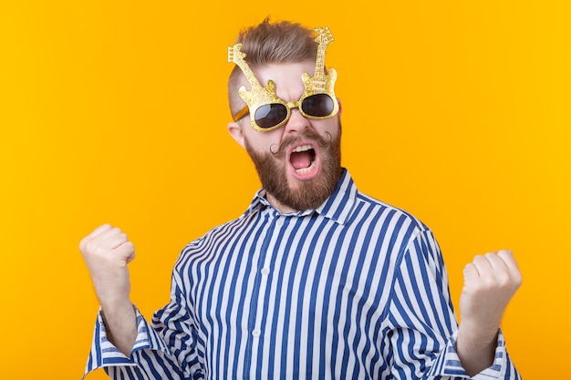 Joven positivo con gafas en forma de guitarras se regocija contra una pared amarilla