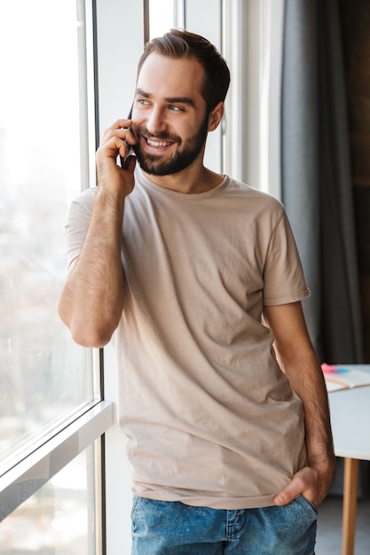 un joven positivo sonriente en el interior de su casa hablando por teléfono móvil.