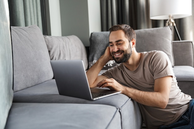 un joven positivo sonriente en el interior de su casa en el sofá usando la computadora portátil.