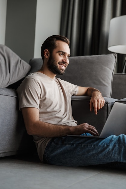 un joven positivo sonriente en el interior de su casa en el sofá usando la computadora portátil.