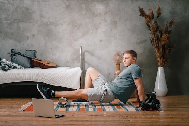 El joven practica deportes en casa. Deportista alegre con cabello rubio estira la pierna en el rodillo de yoga y mira en la computadora portátil en el dormitorio