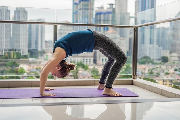 La joven practica yoga por la mañana en su balcón con una vista panorámica de la ciudad y los rascacielos