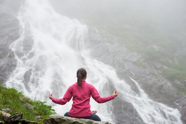 Joven preocupada meditando sobre roca practicando yoga frente a una espectacular cascada