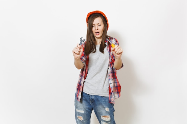 Joven preocupada en ropa casual, casco de construcción protectora con destornillador de juguete y llave aislada sobre fondo blanco. Instrumentos para la renovación de la habitación del apartamento. Reparar el concepto de hogar.