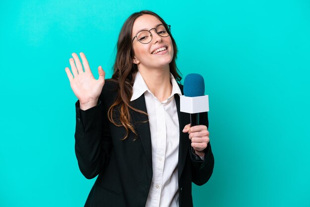 Joven presentadora de televisión aislada de fondo azul saludando con la mano con expresión feliz