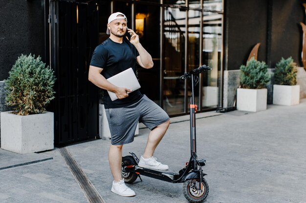 Joven profesional independiente al aire libre Apuesto profesional independiente sosteniendo una computadora portátil y hablando por teléfono en un scooter eléctrico en la calle