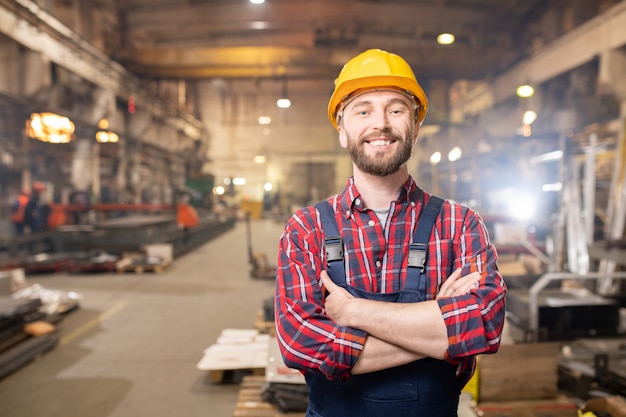 Joven profesional sonriente en monos y casco protector posando dentro de una gran planta industrial