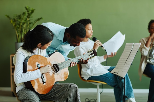 Joven profesor afroamericano de música inclinado sobre colegiala asiática preadolescente con interfaz gráfica de usuario acústica