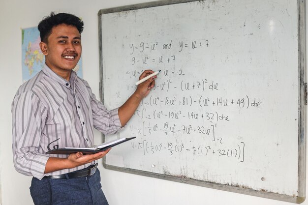 Joven profesor de matemáticas asiático sosteniendo libros y explicando fórmulas matemáticas escritas en la pizarra