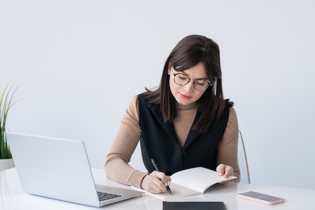 Joven profesora o empresaria elegante haciendo notas de trabajo en el cuaderno mientras está sentado frente a la computadora portátil