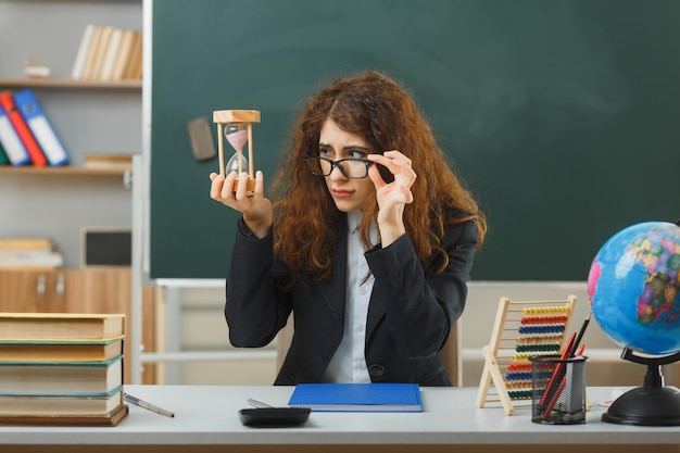 una joven profesora pensante con gafas sosteniendo un reloj de arena sentada en un escritorio con herramientas escolares en el aula