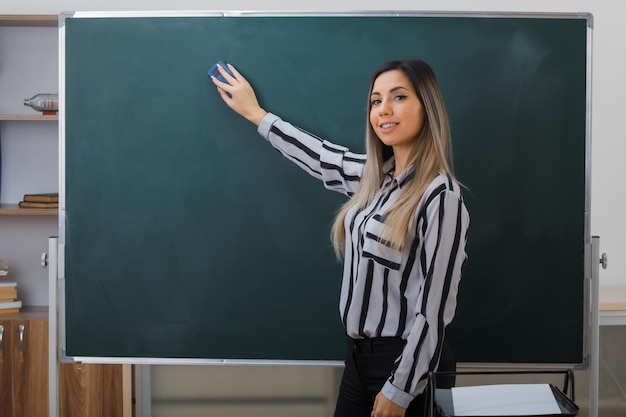 joven profesora de pie cerca de la pizarra en el aula explicando la lección limpiando la pizarra con una esponja sonriendo confiada